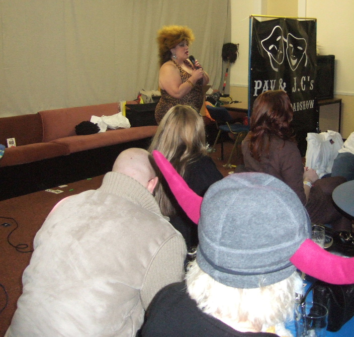 Comedian Christine Coles live on stage at Potters Langley Mill in a leopard print dress with a banner for Pav and JC's Variety Roadshow in the background.